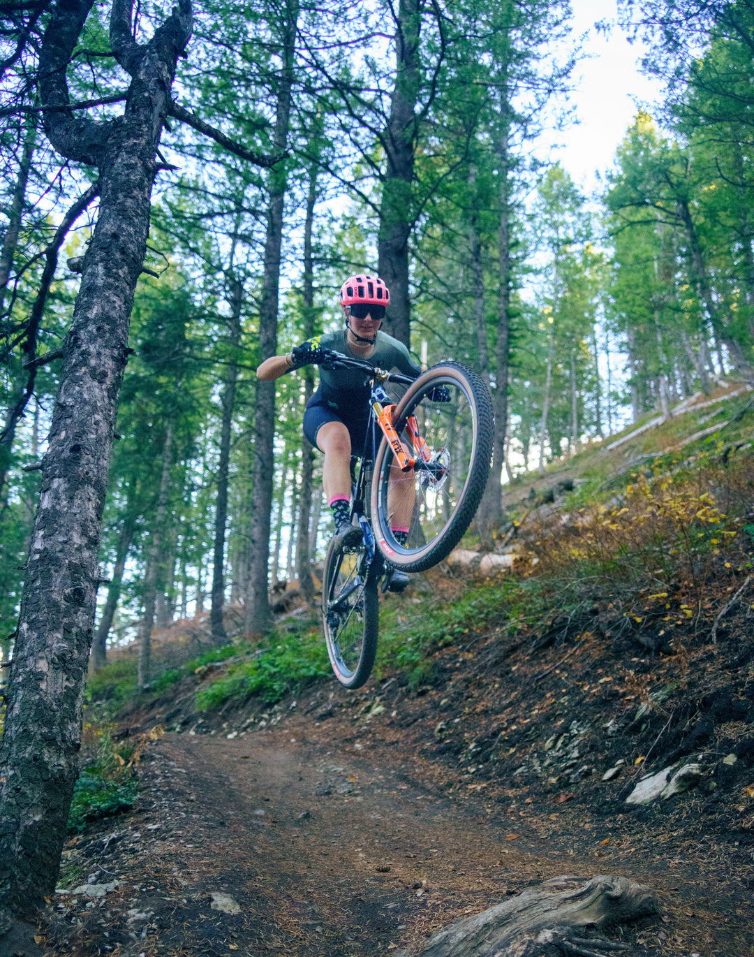 Woman with a pink helmet on a Jibe mountain bike in mid-air over a dirt trail in a forest